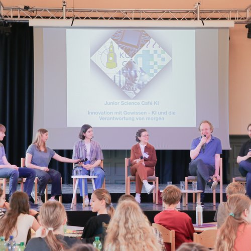 Auf einer Bühne diskutieren Schüler*innen und Wissenschaftler*innen. Sie lächeln. Davor sitzen diverse Schüler*innen.
